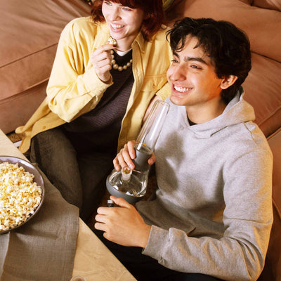 Couple using a clear bong with grey cover while enjoying a movie and a bowl of popcorn. 