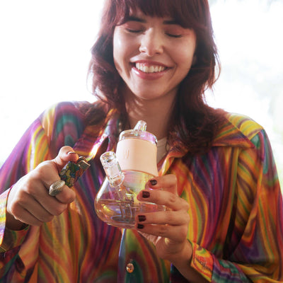 Model laughing while lighting a pink bubbler bong wearing a rainbow shirt. 