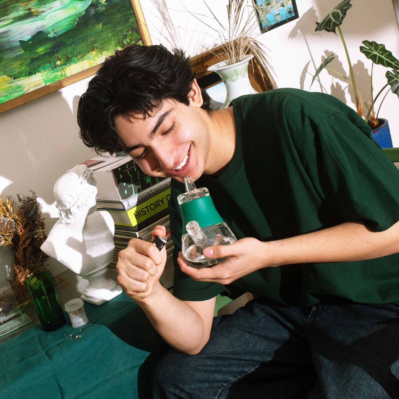 man in a green shirt using a forest green bubbler bong in front of potted plants and books. 