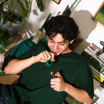 Man using a forest green pipe in front of potted plants and books. 