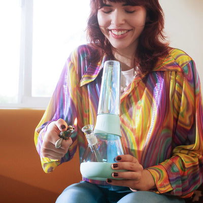 woman in a colorful shirt using a sky blue iridescent bong on an orange sofa. 