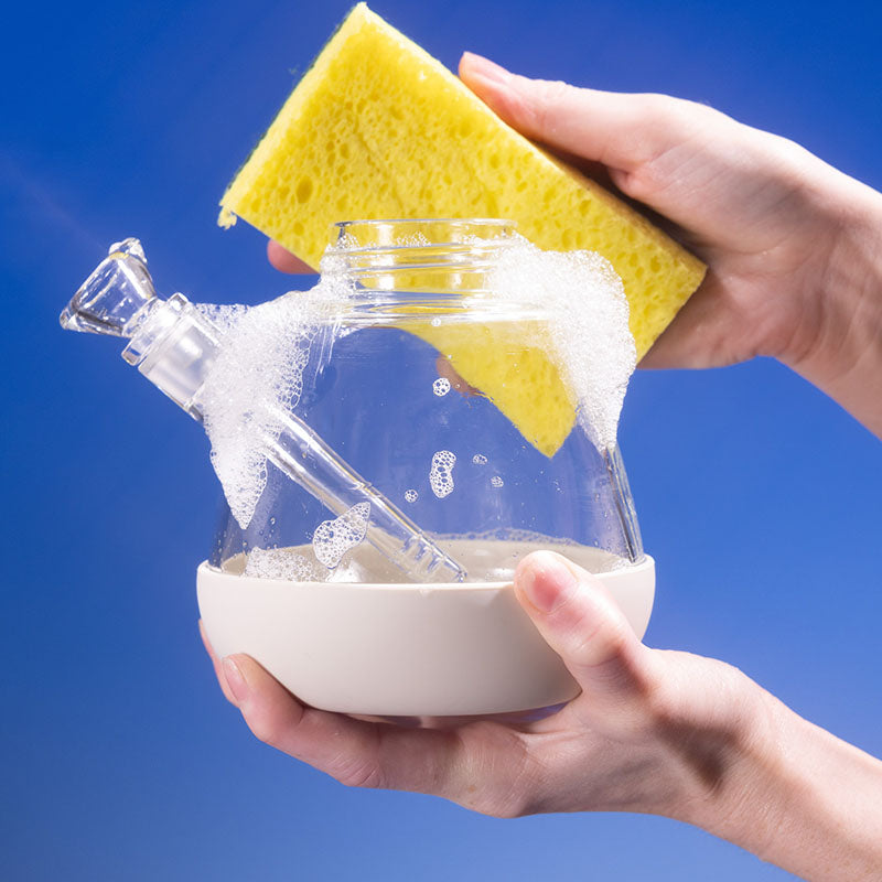 Person cleaning Weeday glass bong base with yellow sponge, white foamy bubbles, against blue background.