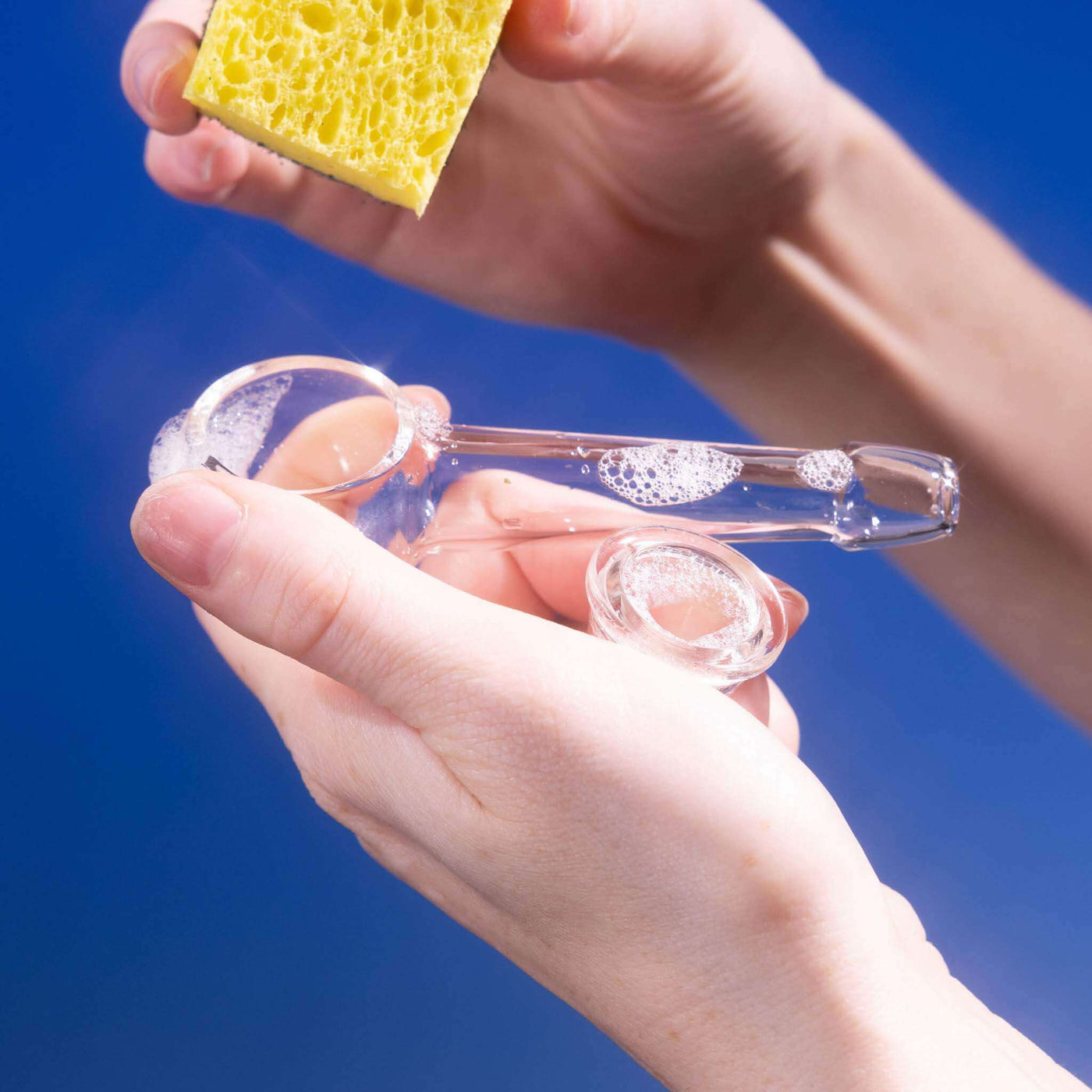 Hand cleaning a Weeday glass pipe with a sponge, highlighting the ease of cleaning, against a blue background.