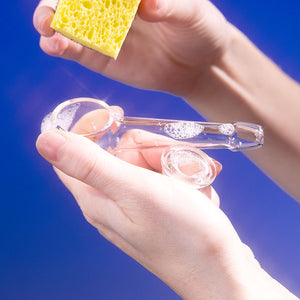 Hand cleaning a Weeday glass pipe with a sponge, highlighting the ease of cleaning, against a blue background.