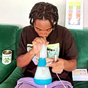 Black male customer in t-shirt using Weeday water pipe on a green sofa, with Weeday package boxes in the background
