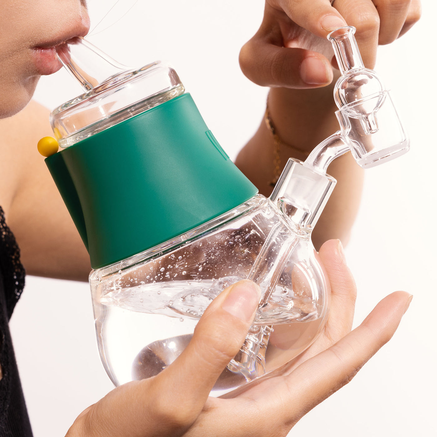 A person is holding a glass dab rig with forest green silicon cover, rotating the cap as water bubbles inside, with a white background