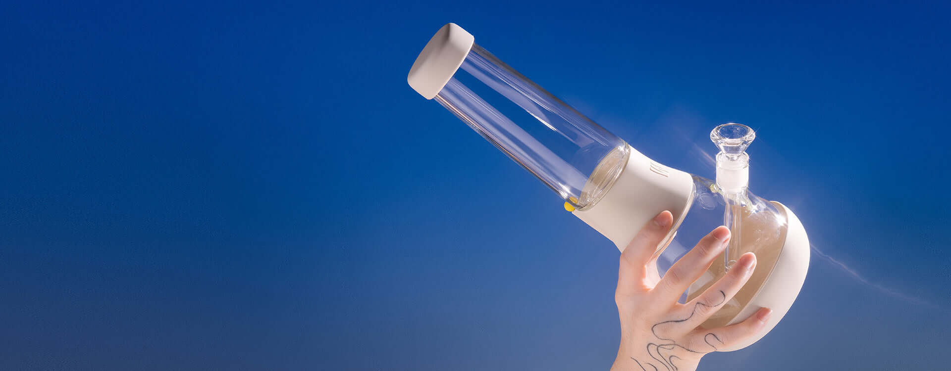 Hand holding up a clear, glowy, crystal-like glass beaker bong with a cream-white silicone cover against a dreamy blue background.
