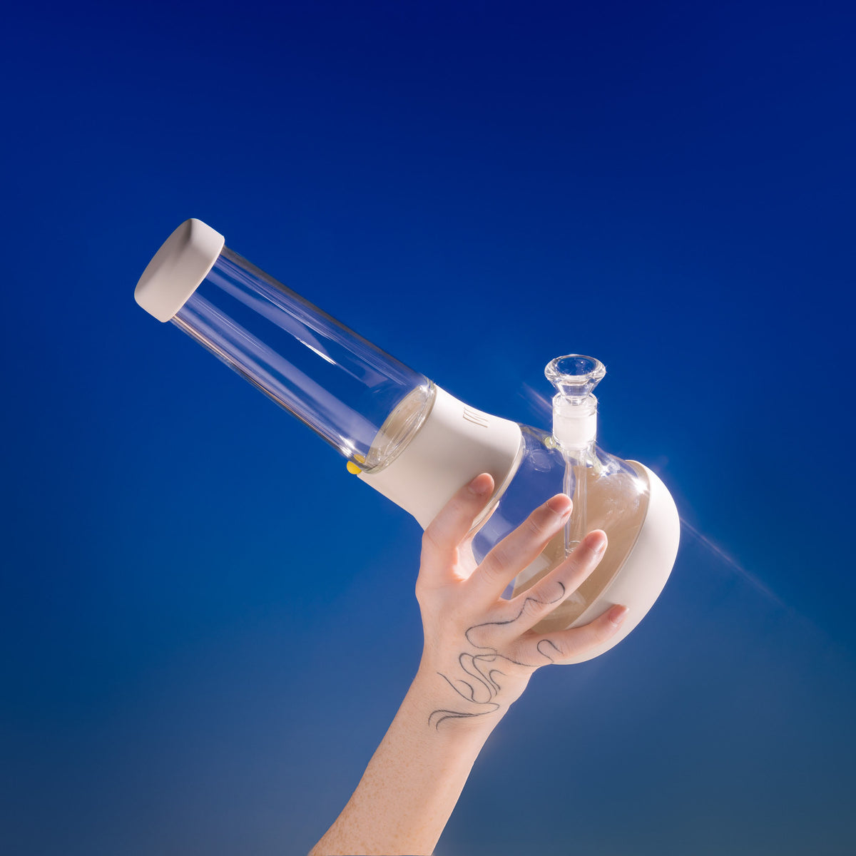 Hand holding up a clear, glowy, crystal-like glass beaker bong with a cream-white silicone cover against a vibrant blue background.