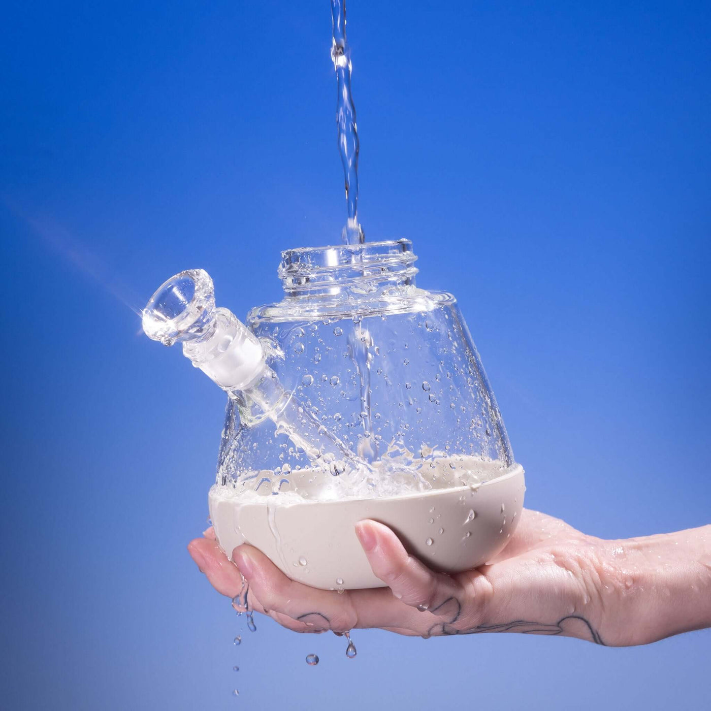 Hand rinsing a detached Weeday beaker bong base with running water, against a blue background.