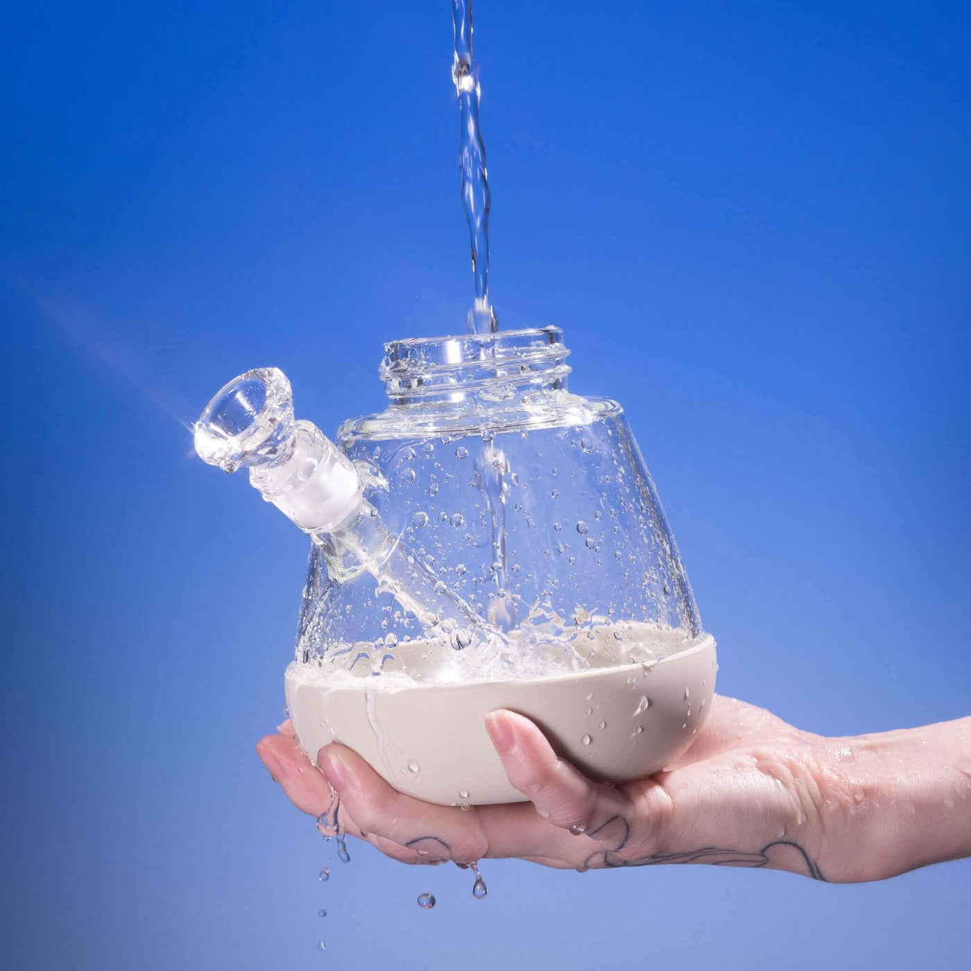 Hand rinsing a detached Weeday beaker bong base with running water, against a blue background.