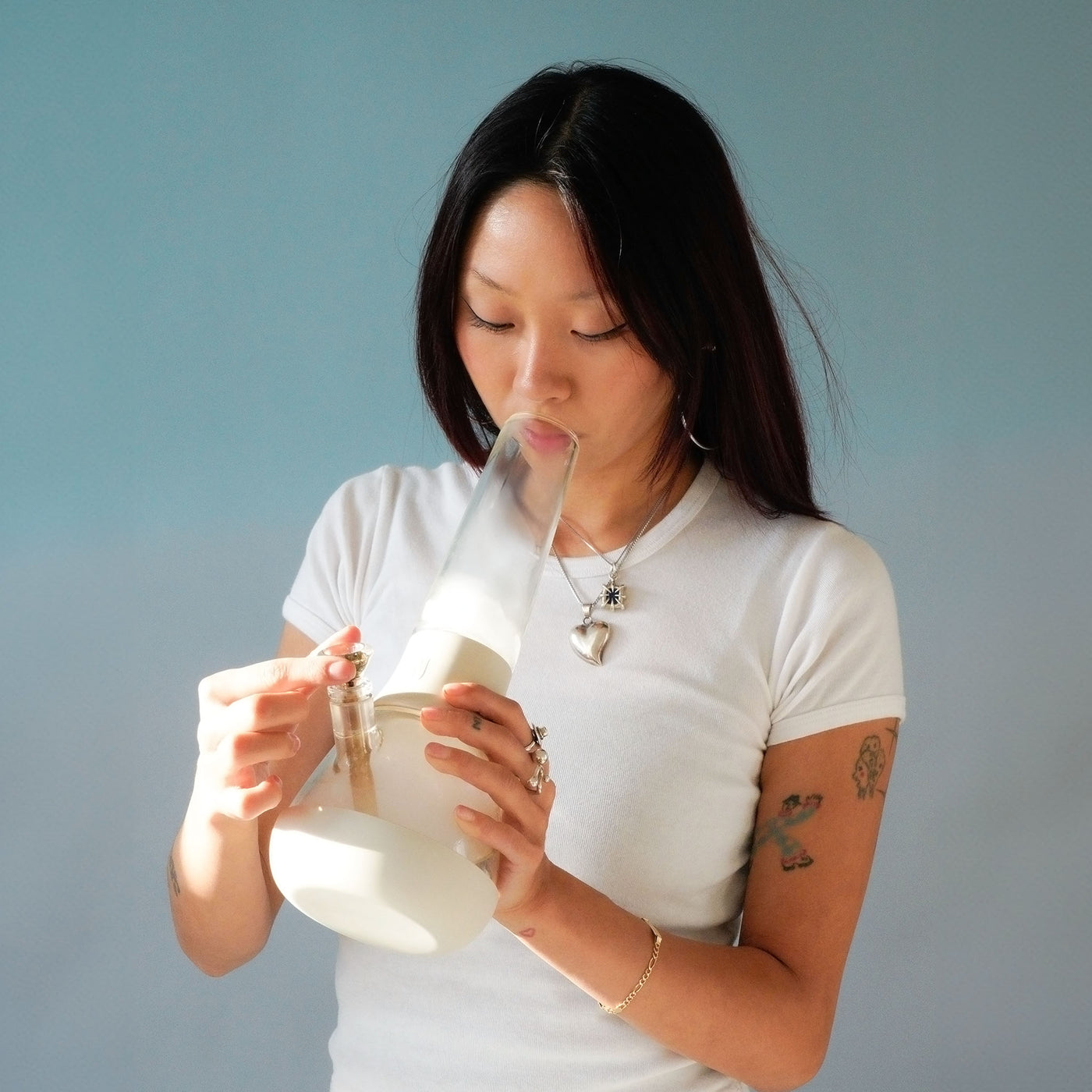 An asian woman smoking with a Weeday glass bong in cream white color, standing in front of a light blue wall.