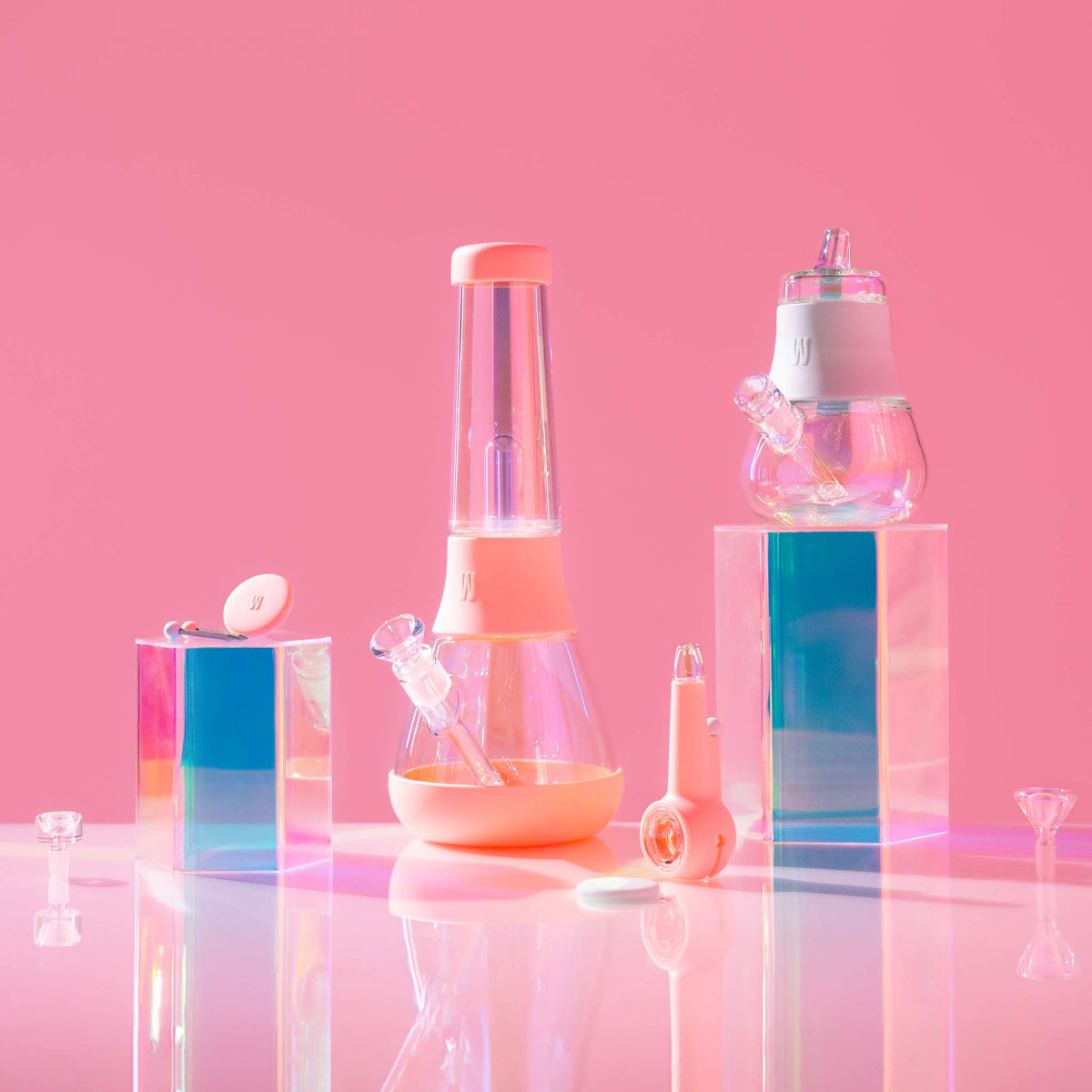 A vibrant collection of iridescent glass bongs, bubblers, and pipes with silicone grips, set against a pink reflective background and acrylic glass box.