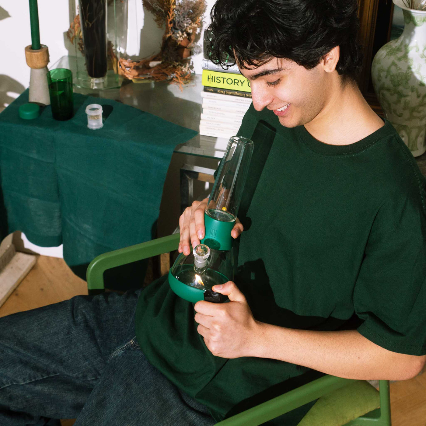 A model sitting on a green chair, lighting a forest-green water bong, surrounded by books and decor, creating a cozy and relaxed vibe.