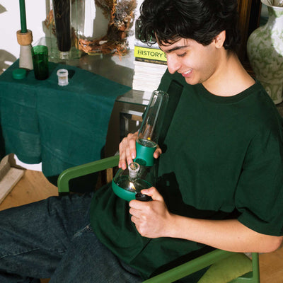 A model sitting on a green chair, lighting a forest-green water bong, surrounded by books and decor, creating a cozy and relaxed vibe.