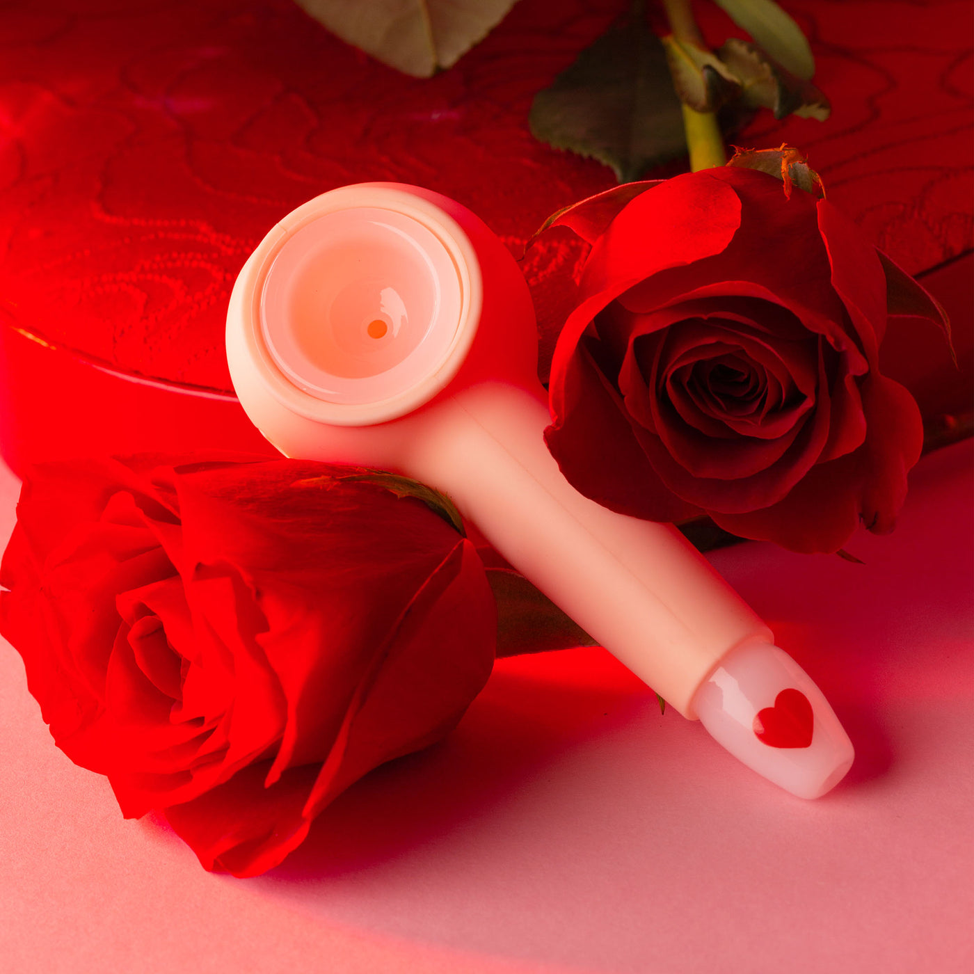 Pink smoking pipe with what base and heart decal on mouth piece. Pipe is laying on two red roses with a red heart chocolate box. 