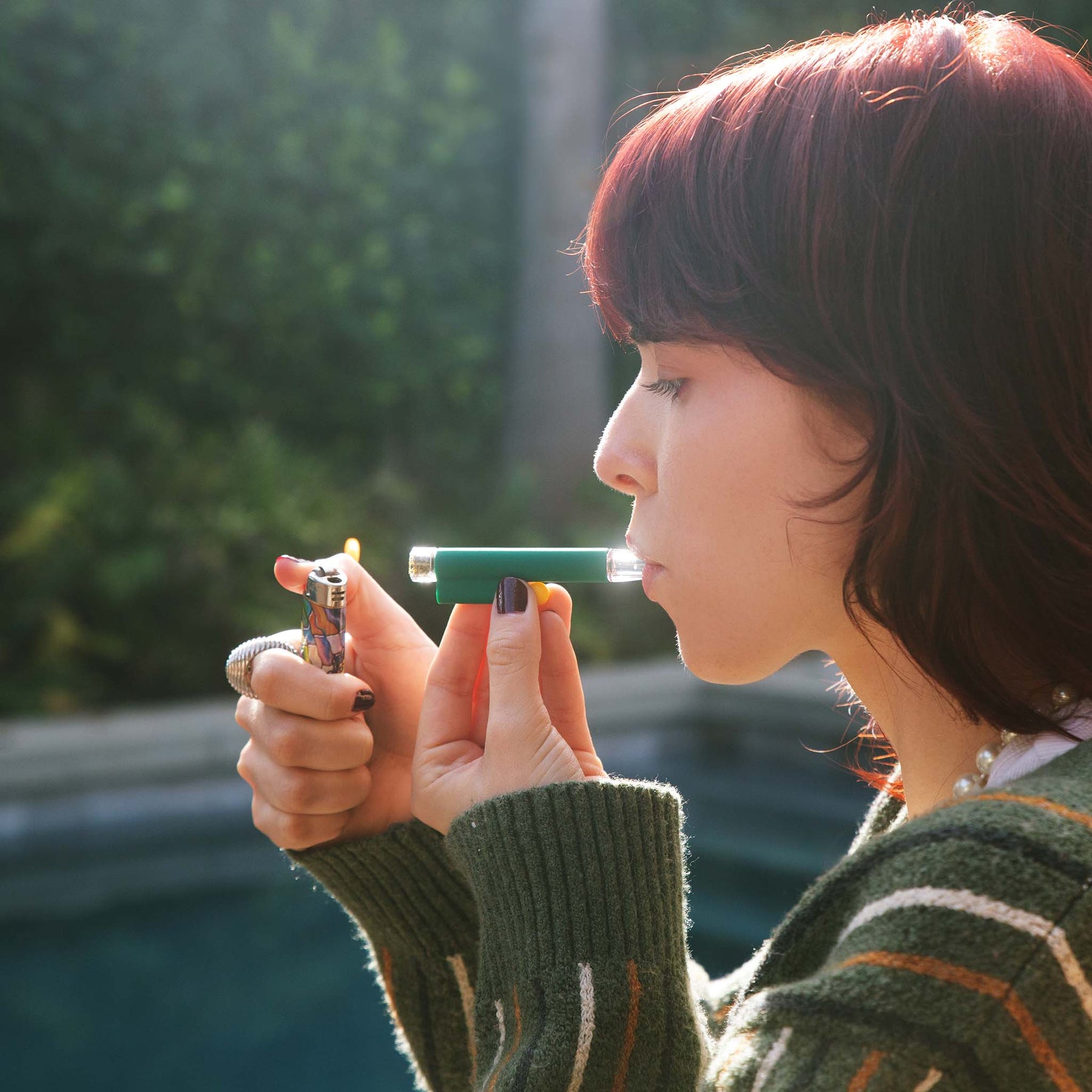 Model smoking with a forest green one-hitter pipe with a yellow accent, enjoying a smooth hit outdoors near a pool.