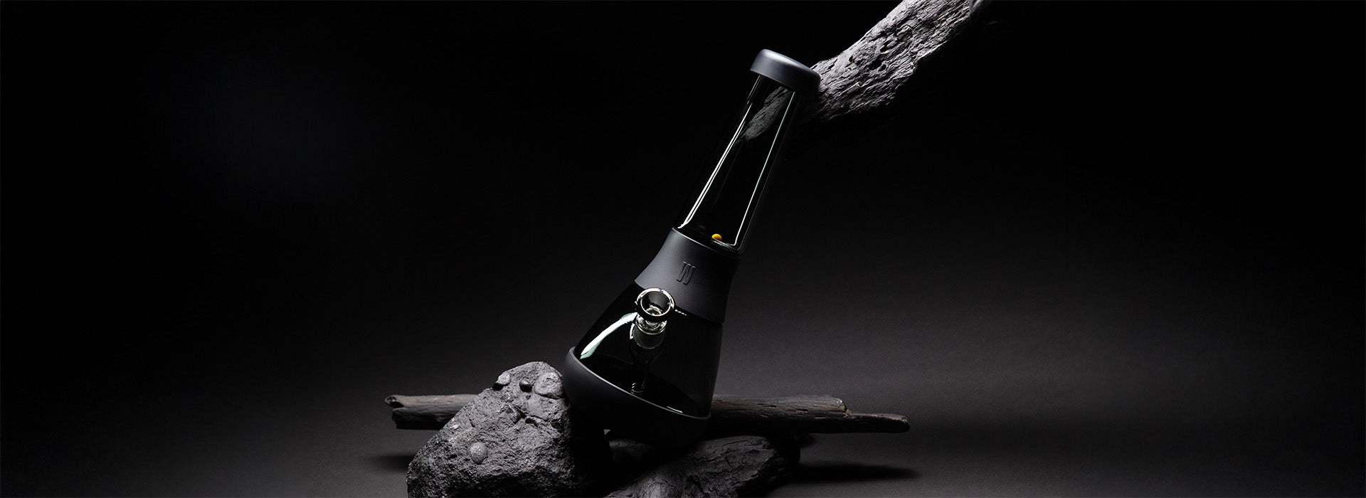 A tilted Weeday black glass bong displayed on rugged stones and driftwood against a dark studio background