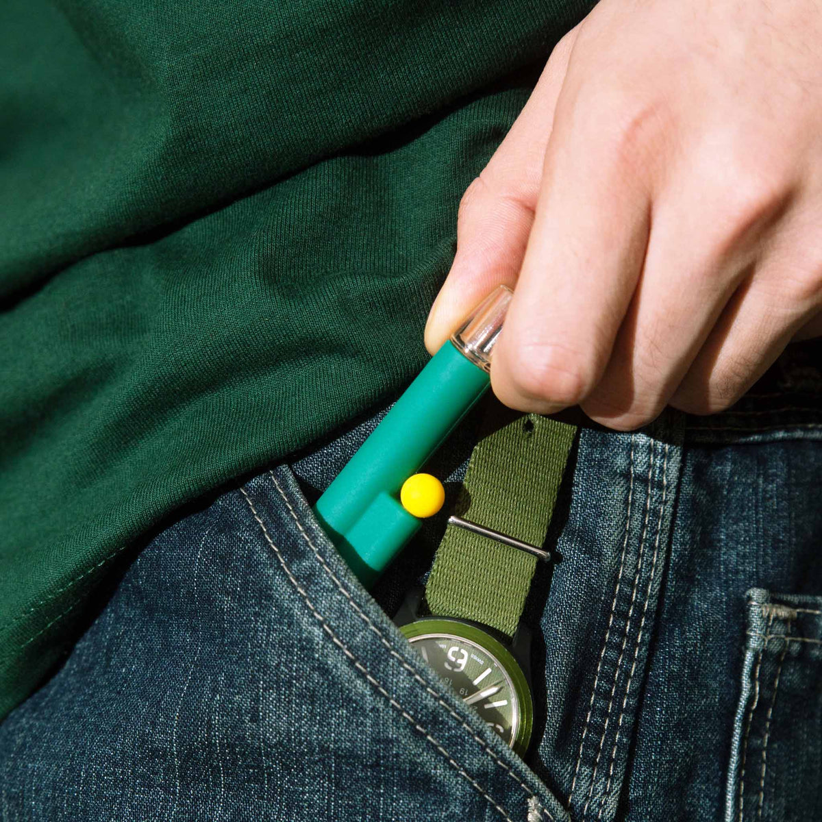 Travel-friendly one-hitter pipe conveniently fitting into a jeans pocket next to a green watch strap.