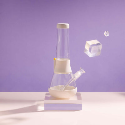 Product photo of sleek glass bong on cream white cover in a purple setting, with glowy acrylic glass beside it.