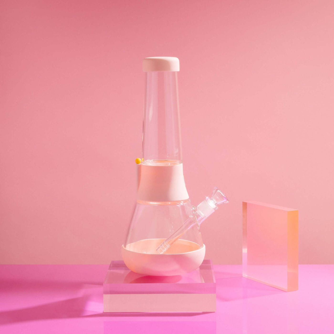 Product photo of sleek glass bong on baby pink cover in a pink setting, with glowy acrylic glass beside it.