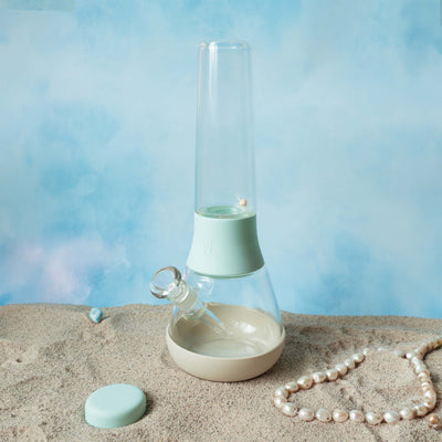 Side view of the Weeday custom bong in Sky and cream mix on sandy surface with pearls, against a cloudy sky backdrop.