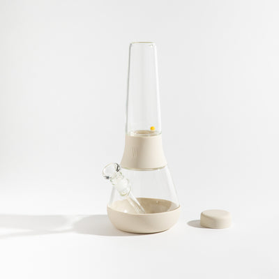 Product photo of a glass waterpipe with cream white silicone covers, cap uncovered, placed on a table.