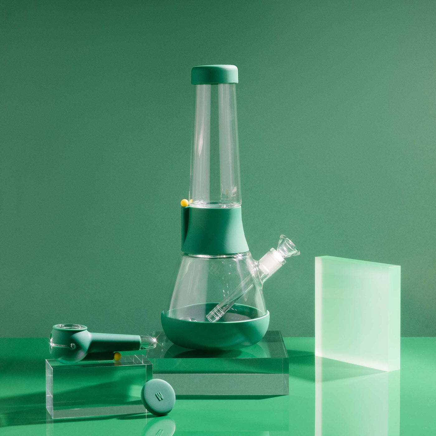  A bundle set of the Weeday beaker bong and silicone pipe with glass bowl, sitting on acrylic blocks, showcased in a forest green studio setting.