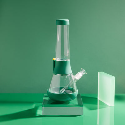 Product photo of sleek glass bong on forest green cover in a green setting, with glowy acrylic glass beside it.