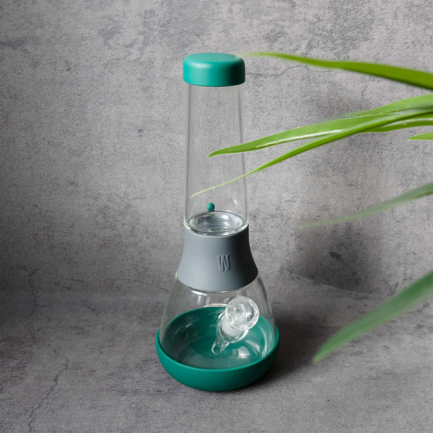 Front view of Weeday glass bong: green base & cap, gray connector, on a textured backdrop with foreground leaves.