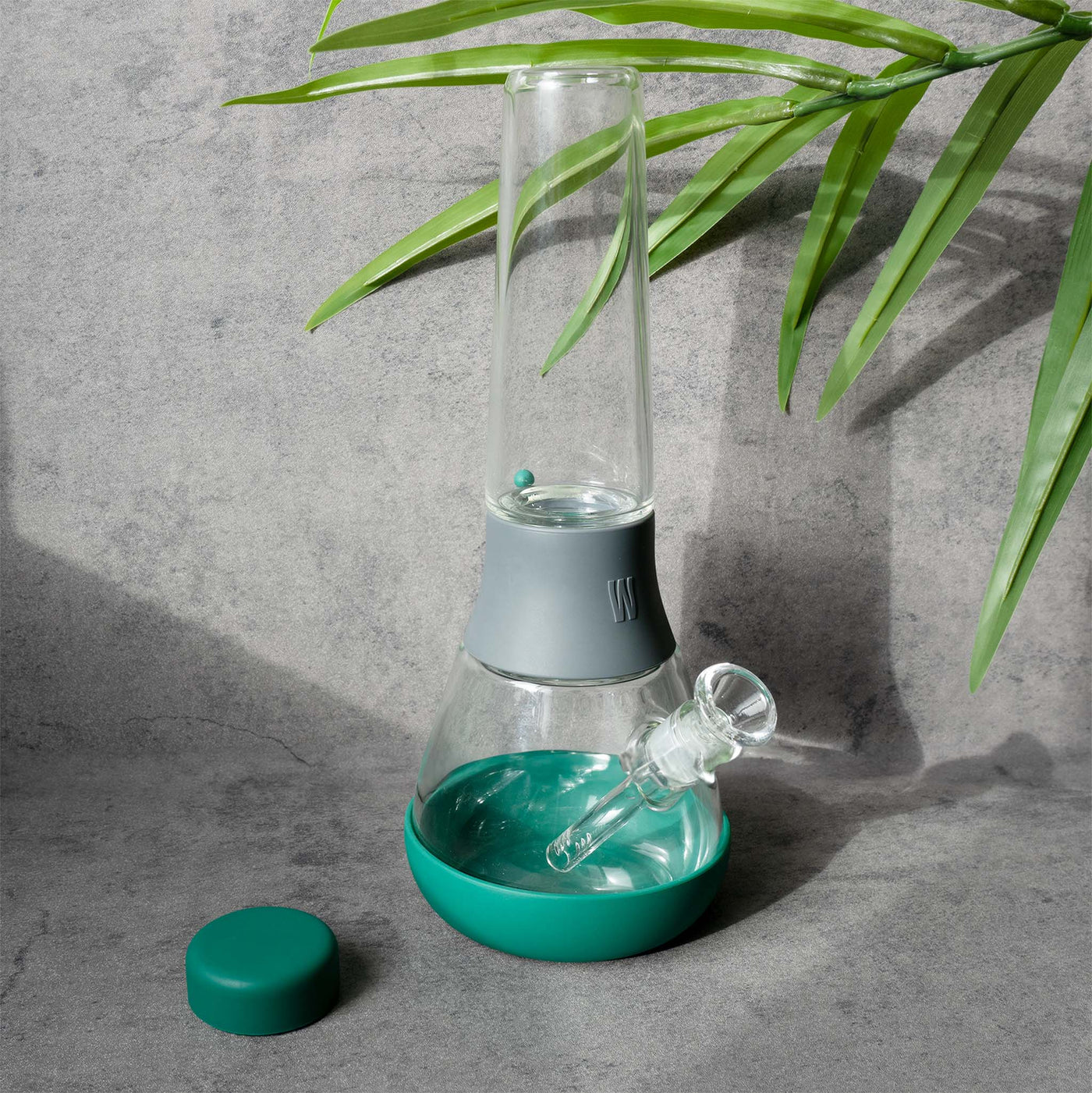 Front view of Weeday glass bong: green base & cap, gray connector, on a textured backdrop with foreground leaves and cap aside. 