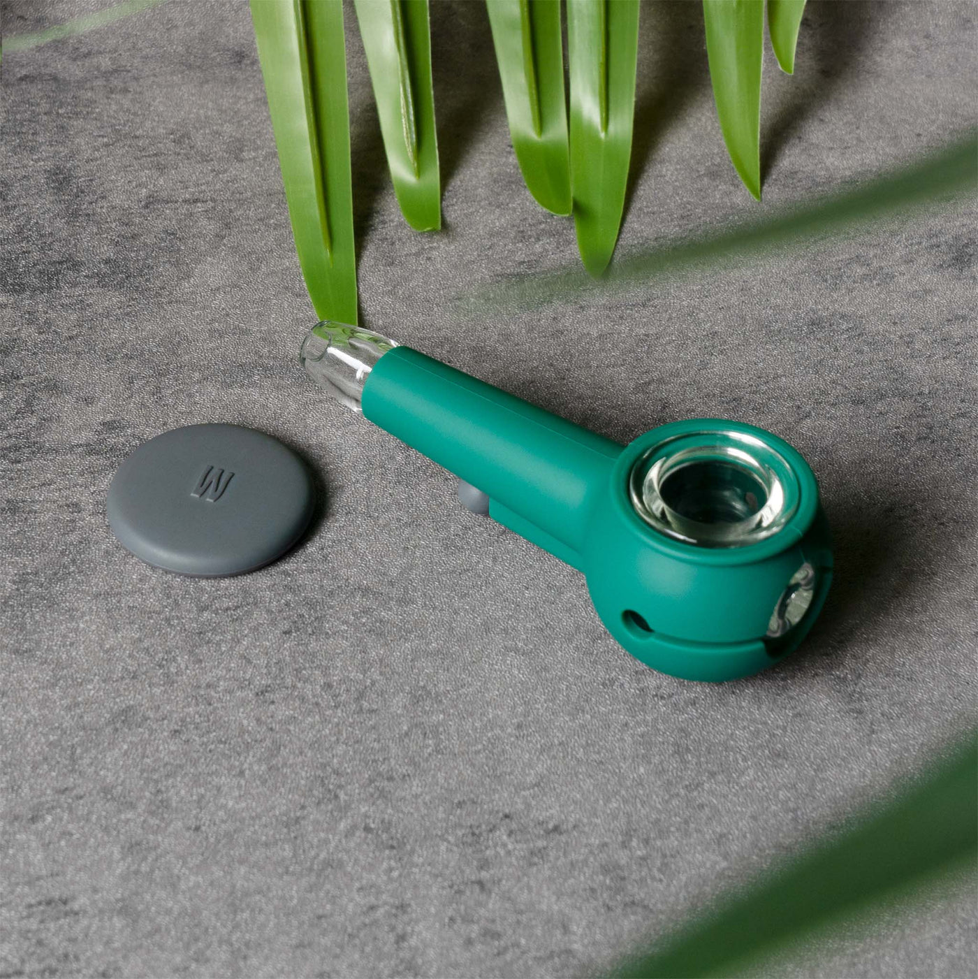 Forest green silicone pipe with a smoke gray lid and clear glass bowl, set against a textured background and fresh green leaves.