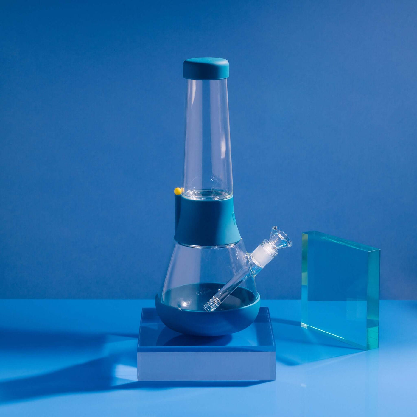 Product photo of sleek glass bong on midnight blue cover in a blue setting, with glowy acrylic glass beside it.