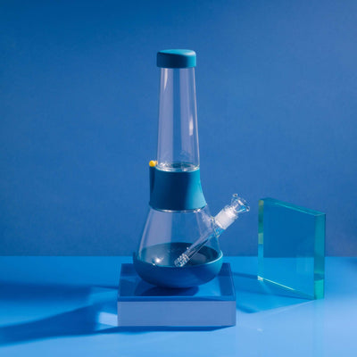 Product photo of sleek glass bong on midnight blue cover in a blue setting, with glowy acrylic glass beside it.
