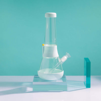 Product photo of sleek glass bong on sky blue cover in a mint setting, with glowy acrylic glass beside it.