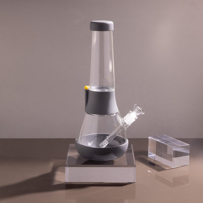 Product photo of sleek glass bong on smoke gray cover in a gray setting, with glowy acrylic glass beside it.