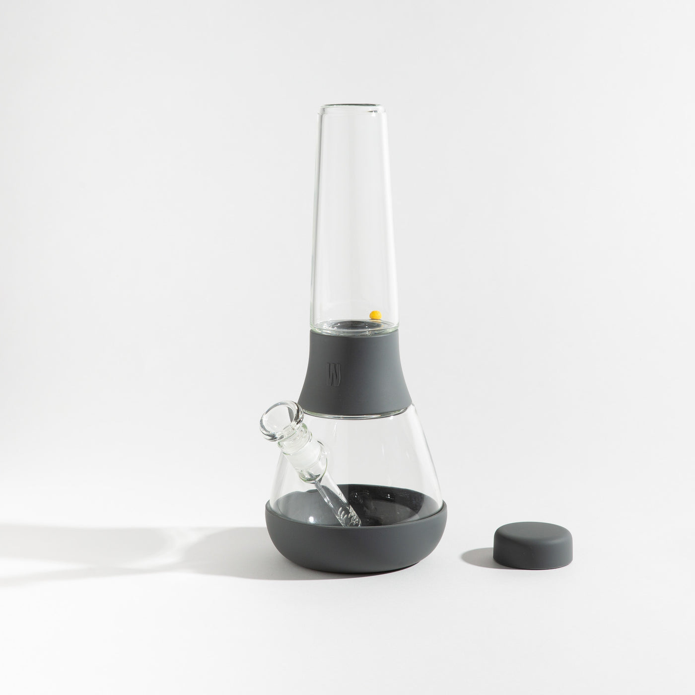 Product photo of a glass waterpipe with smoke gray silicone covers, cap uncovered, placed on a table.